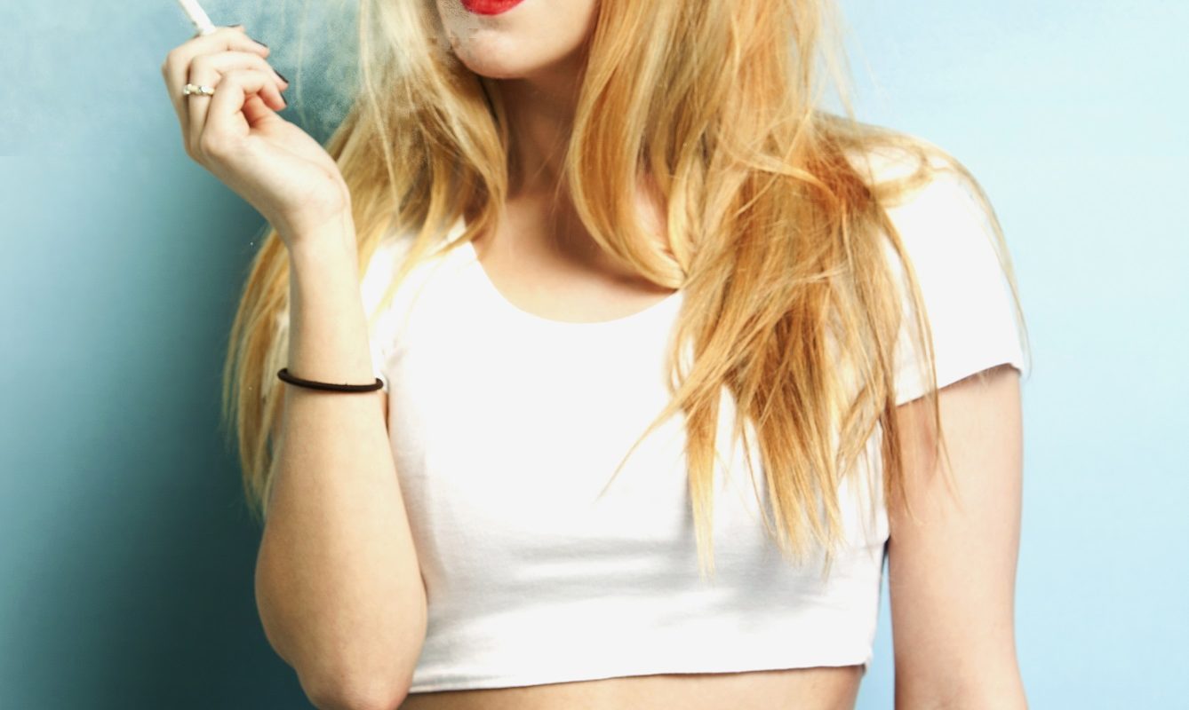 Portrait of young blond woman holding cigarette against blue background